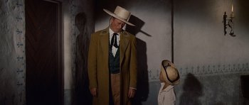 Movie still from “The Alamo” (1960), directed by John Wayne – A man wearing a hat and a long coat standing next to a child; Medium shot, Low angle