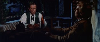 Movie still from “The Alamo” (1960), directed by John Wayne – An older man sitting at a table with a bottle of wine; Medium shot, Over the shoulder angle