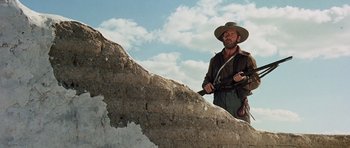 Movie still from “The Alamo” (1960), directed by John Wayne – A man holding a gun standing on top of a rock wall; Medium shot, Low angle