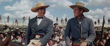 Movie still from “The Alamo” (1960), directed by John Wayne – Two men in cowboy hats are standing next to each other; Medium shot, Low angle