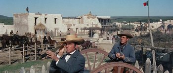 Movie still from “The Alamo” (1960), directed by John Wayne – Two men sitting on a bench in front of some buildings; Medium shot, Over the shoulder angle