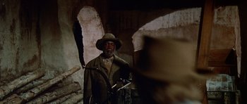 Movie still from “The Alamo” (1960), directed by John Wayne – A man in a hat and a jacket standing next to a wall; Medium shot, Over the shoulder angle