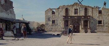 Movie still from “The Alamo” (1960), directed by John Wayne – A man in a cowboy hat is walking in front of an old stone building; Wide shot, Low angle