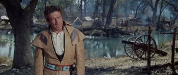 Movie still from “The Alamo” (1960), directed by John Wayne – A man in a leather jacket standing next to a body of water; Medium shot, Over the shoulder angle
