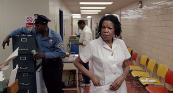 Movie still from “The Ambulance” (1990), directed by Larry Cohen – An older woman in a white lab coat is standing in a room; Medium shot, Over the shoulder angle