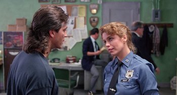 Movie still from “The Ambulance” (1990), directed by Larry Cohen – A police officer talking to a man in a room with other people; Medium shot, Over the shoulder angle