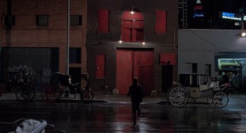 Movie still from “The Ambulance” (1990), directed by Larry Cohen – A person walking in the rain in front of a red building; Extreme Wide shot, Over the shoulder angle
