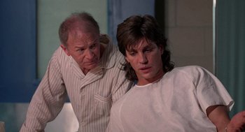 Movie still from “The Ambulance” (1990), directed by Larry Cohen – A man standing next to another man in a room; Medium shot, Over the shoulder angle
