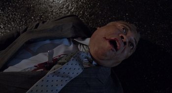 Movie still from “The Ambulance” (1990), directed by Larry Cohen – A man in a suit and tie laying on the ground with a knife in his mouth; Close Up shot, Overhead angle