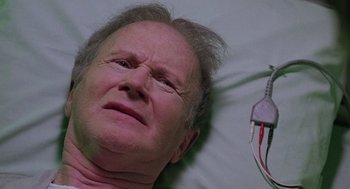 Movie still from “The Ambulance” (1990), directed by Larry Cohen – An older man laying in a hospital bed with an electrical device hanging from the side of his head; Close Up shot, High angle