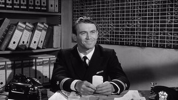 Movie still from “The Americanization of Emily” (1964), directed by Arthur Hiller – A man sitting at a table holding a card; Medium shot, Over the shoulder angle