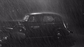 Movie still from “The Americanization of Emily” (1964), directed by Arthur Hiller – An old car is driving in the rain on the street; Wide shot, High angle