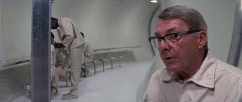 Movie still from “The Andromeda Strain” (1971), directed by Robert Wise – An older man wearing glasses in a room with benches; Medium shot, Over the shoulder angle