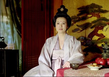 Movie still from “The Assassin” (2015), directed by Hsiao-Hsien Hou – A woman in a traditional asian dress sitting at a table; Medium shot, Low angle