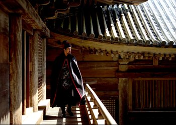 Movie still from “The Assassin” (2015), directed by Hsiao-Hsien Hou – A man wearing a black coat and a black hat standing on a wooden railing outside of a building; Wide shot, Low angle