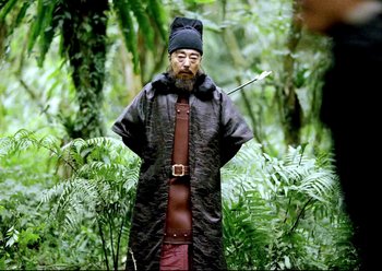 Movie still from “The Assassin” (2015), directed by Hsiao-Hsien Hou – A man in a black coat and a hat holding a bow and arrow in the woods; Medium shot, Low angle
