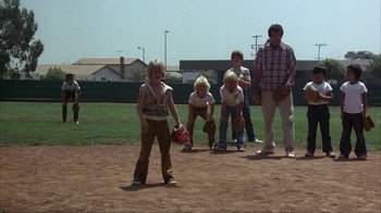 Movie still from “The Bad News Bears” (1976), directed by Michael Ritchie – A group of young people standing on top of a baseball field; Wide shot, Over the shoulder angle