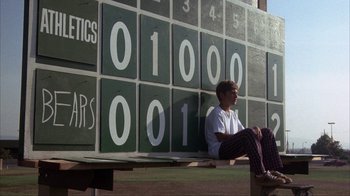 Movie still from “The Bad News Bears” (1976), directed by Michael Ritchie – A man sitting on a bench in front of a scoreboard; Wide shot, Low angle