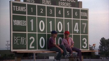 Movie still from “The Bad News Bears” (1976), directed by Michael Ritchie – Two men sitting on top of a bench in front of a scoreboard; Wide shot, Low angle