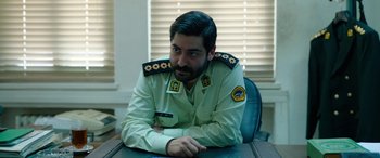 Movie still from “Holy Spider” (2022), directed by Ali Abbasi – A man sitting at a table wearing a military uniform; Medium shot, Over the shoulder angle
