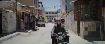 Movie still from “Holy Spider” (2022), directed by Ali Abbasi – A man riding a motorcycle down a street with a man on the back of it; Wide shot, Over the shoulder angle