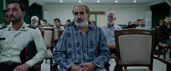 Movie still from “Holy Spider” (2022), directed by Ali Abbasi – An older man sitting in front of a group of older men; Medium shot, Over the shoulder angle