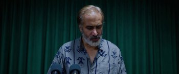 Movie still from “Holy Spider” (2022), directed by Ali Abbasi – A man with a gray beard is standing in front of microphones; Close Up shot, High angle