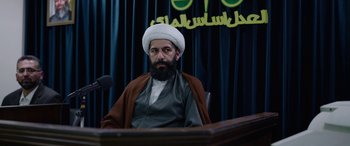 Movie still from “Holy Spider” (2022), directed by Ali Abbasi – A man with a beard and a white hat sitting in front of a microphone; Medium shot, Low angle