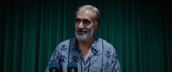 Movie still from “Holy Spider” (2022), directed by Ali Abbasi – A man with a gray beard is standing in front of microphones; Close Up shot, Over the shoulder angle