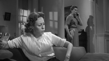 Movie still from “The Bad Seed” (1956), directed by Mervyn LeRoy – A woman sitting on a couch in front of another woman on a phone; Medium shot, Low angle