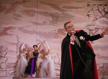 Movie still from “The Band Wagon” (1953), directed by Vincente Minnelli – A man wearing a cape and holding a microphone in front of a group of women; Wide shot, Low angle