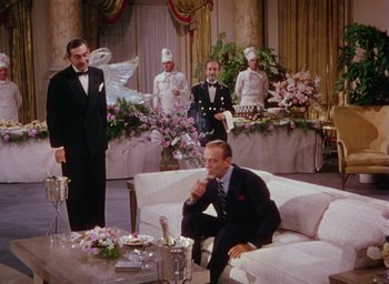 Movie still from “The Band Wagon” (1953), directed by Vincente Minnelli – A man sitting on top of a white couch next to flowers; Wide shot, High angle