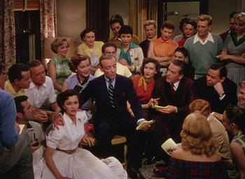 Movie still from “The Band Wagon” (1953), directed by Vincente Minnelli – A group of people gathered around a man in a suit; Wide shot, High angle