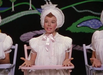Movie still from “The Band Wagon” (1953), directed by Vincente Minnelli – A woman in a white dress and a white hat is sitting in a pink chair; Medium shot, High angle