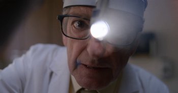 Movie still from “Horse Girl” (2020), directed by Jeff Baena – An older man wearing a white lab coat and a white hat with a magnifying glass on it's head; Close Up shot, Low angle