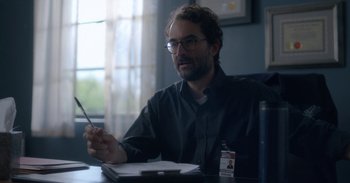 Movie still from “Horse Girl” (2020), directed by Jeff Baena – A man sitting at a table holding a pen; Close Up shot, Over the shoulder angle