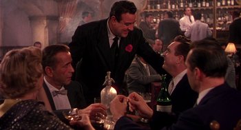 Movie still from “The Barefoot Contessa” (1954), directed by Joseph L. Mankiewicz – A group of people sitting at a table with drinks; Medium shot, Over the shoulder angle