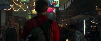 Movie still from “The Beach” (2000), directed by Danny Boyle – A man in a red shirt is standing in the street with a backpack; Wide shot, Over the shoulder angle