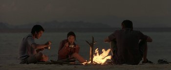 Movie still from “The Beach” (2000), directed by Danny Boyle – A woman sitting on the ground next to a fire; Wide shot, Over the shoulder angle