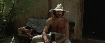 Movie still from “The Beach” (2000), directed by Danny Boyle – A shirtless man sitting on a couch wearing a straw hat; Medium shot, Low angle