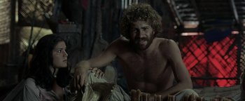 Movie still from “The Beach” (2000), directed by Danny Boyle – A shirtless man sitting on top of a wooden bench; Medium shot, Low angle