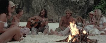 Movie still from “The Beach” (2000), directed by Danny Boyle – A couple of men sitting on top of a sandy beach next to a fire; Wide shot, Low angle