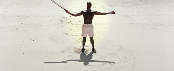 Movie still from “The Beach” (2000), directed by Danny Boyle – A man standing on a beach holding a baseball bat; Wide shot, Overhead angle