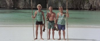 Movie still from “The Beach” (2000), directed by Danny Boyle – Three young men standing on the beach holding skateboards; Wide shot, Low angle