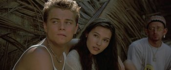 Movie still from “The Beach” (2000), directed by Danny Boyle – A man and a woman sitting next to each other in front of a palm tree; Close Up shot, Low angle
