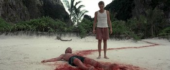 Movie still from “The Beach” (2000), directed by Danny Boyle – A man standing next to another man laying on the beach; Wide shot, Low angle