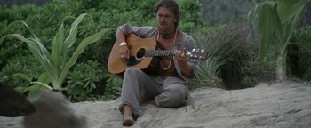 Movie still from “The Beach” (2000), directed by Danny Boyle – A man sitting on the ground playing a guitar; Medium shot, High angle