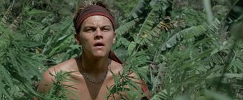 Movie still from “The Beach” (2000), directed by Danny Boyle – A man with a bandana around his head looking at the camera while standing in the grass; Close Up shot, Low angle