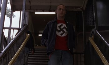 Movie still from “The Believer” (2001), directed by Henry Bean – A man in a red shirt and a black jacket standing on some stairs; Medium shot, Low angle