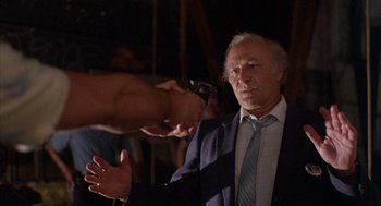 Movie still from “The Believers” (1987), directed by John Schlesinger – An older man in a suit and tie holding a gun; Medium shot, Over the shoulder angle